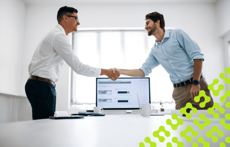 Two business professionals shaking hands across a desk in a bright office, with a computer screen in the background.