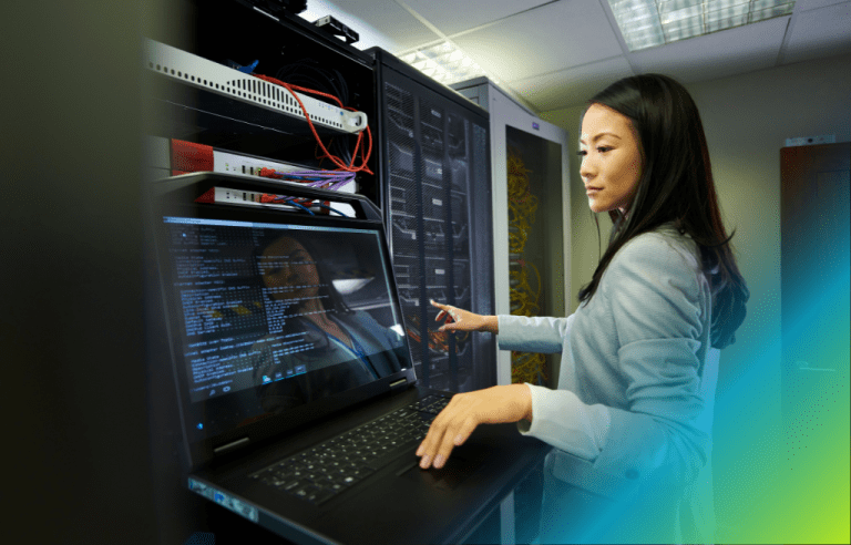 Woman in a server room working on a laptop next to server racks.
