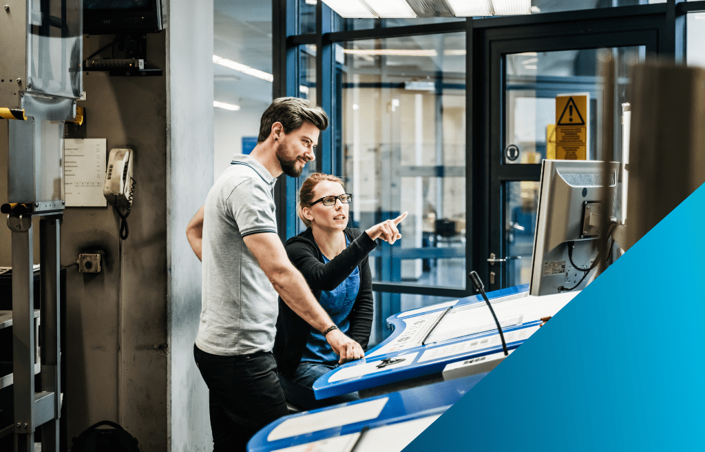 A female technician points to a screen at a control panel while a male supervisor stands next to her, collaborating on an operation in a modern industrial facility