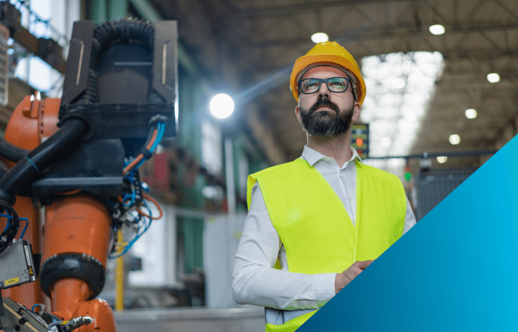 A male engineer in a yellow hard hat and safety vest stands in a factory, with a large orange robotic arm beside him.