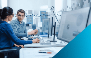 Two professionals, a man and a woman, are working together at a desk with multiple computer monitors in a bright, modern office.