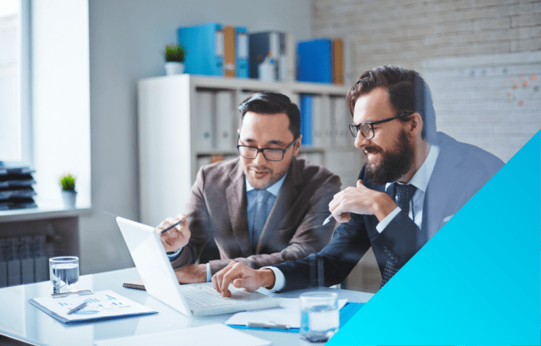 Two men in business suits are seated at a desk, looking at a laptop and discussing work, pointing at the screen with pens.