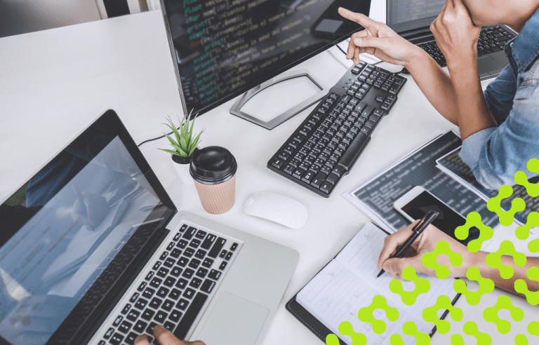 Two individuals collaborate at an office workstation, using a laptop and a desktop monitor displaying code while taking notes in a notebook.