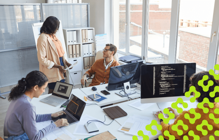 A team of four professionals are working in an office, with two individuals working on laptops, one is focused on a monitor displaying code, and a fourth is leading a discussion near a whiteboard.