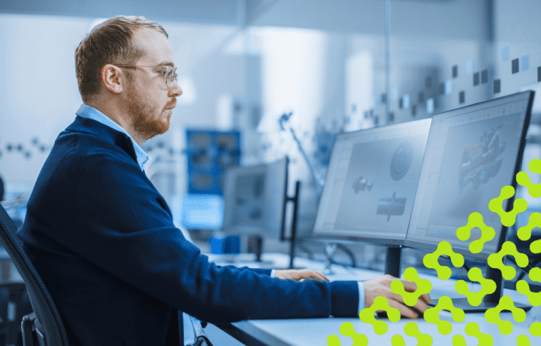A man sits at a workstation with two monitors displaying 3D Computer-Aided Design models of a component.