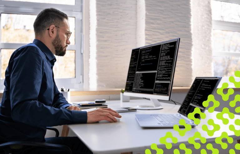 A man sits at an office desk, focusing on code displayed across two screens.