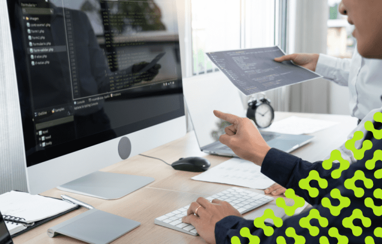 Two professionals review code displayed on a monitor and a document at a workstation, working on a software application.