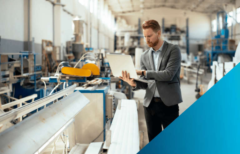 Ma business suit holding a laptop while standing on a factory floor next to a production line with industrial machinery.