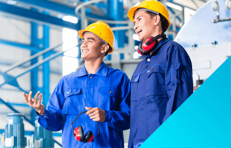 Two smiling factory workers in yellow hard hats and blue uniforms stand together.