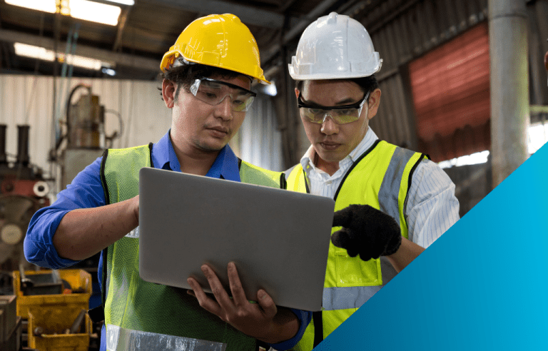 Two industrial workers in hard hats and safety gear discussing data on a laptop.