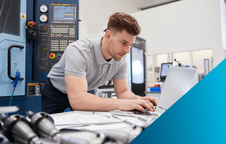 Man typing on a laptop in a room full of technical controls and parts.